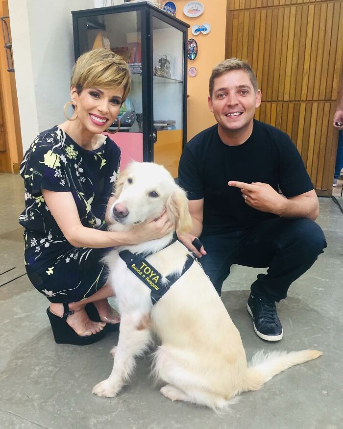 Two people smiling while posing with a golden retriever dog wearing a harness indoors, symbolizing human-animal bonding.