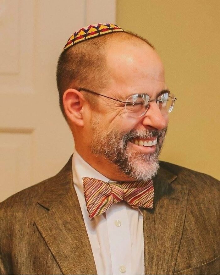 Man wearing glasses and a patterned bow tie smiling, related to Dr Robby in The Pitt real doctor discussion.