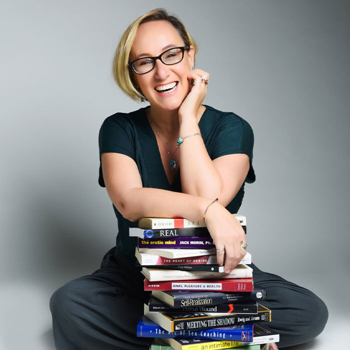 Smiling woman with glasses sitting cross-legged, leaning on a stack of books about intimacy and sexuality topics.