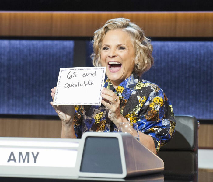 Amy Sedaris holding a sign and smiling during a TV broadcast, related to her transphobic comments on CNN New Year's Eve.