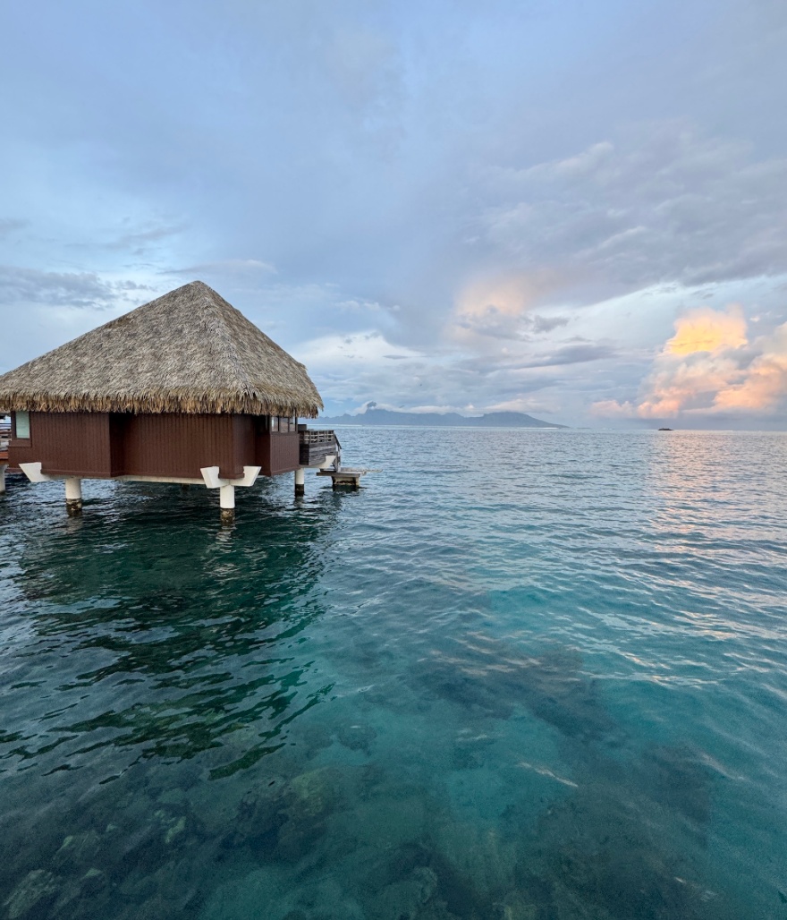Overwater bungalow surrounded by vibrant shades of blue water in French Polynesia at sunset.