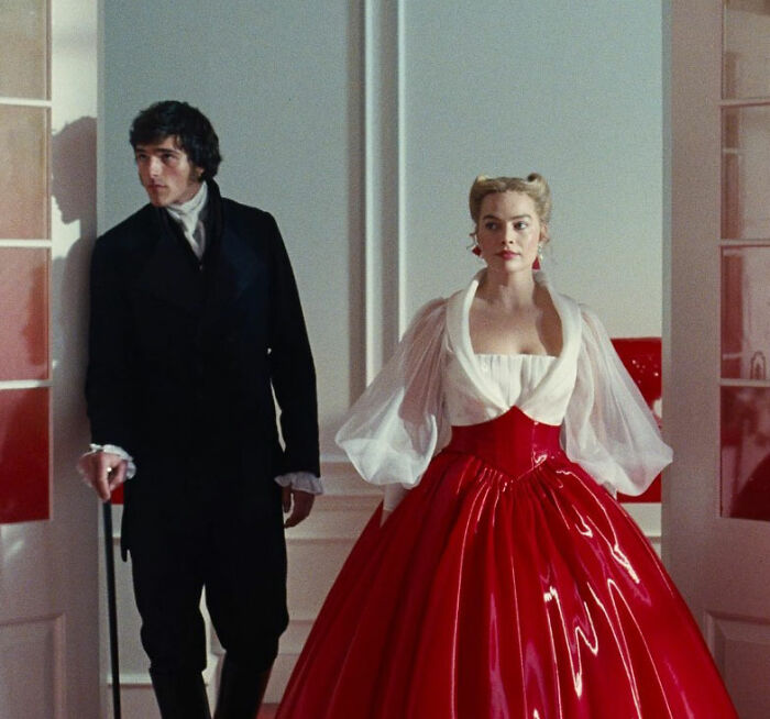 Woman in a red bathing suit with sleeves and a voluminous skirt standing next to a man in period costume indoors.