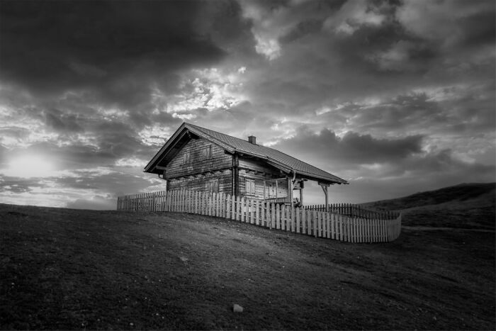 Black and white photo of a wooden cabin on a hill under dramatic cloudy skies, showcasing stunning animal photos theme.