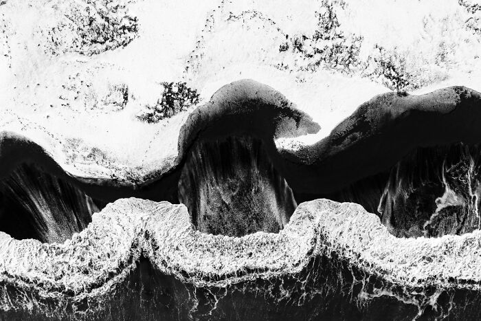 Aerial black and white photo capturing dramatic ocean waves crashing along a rugged shoreline with textured sand patterns.
