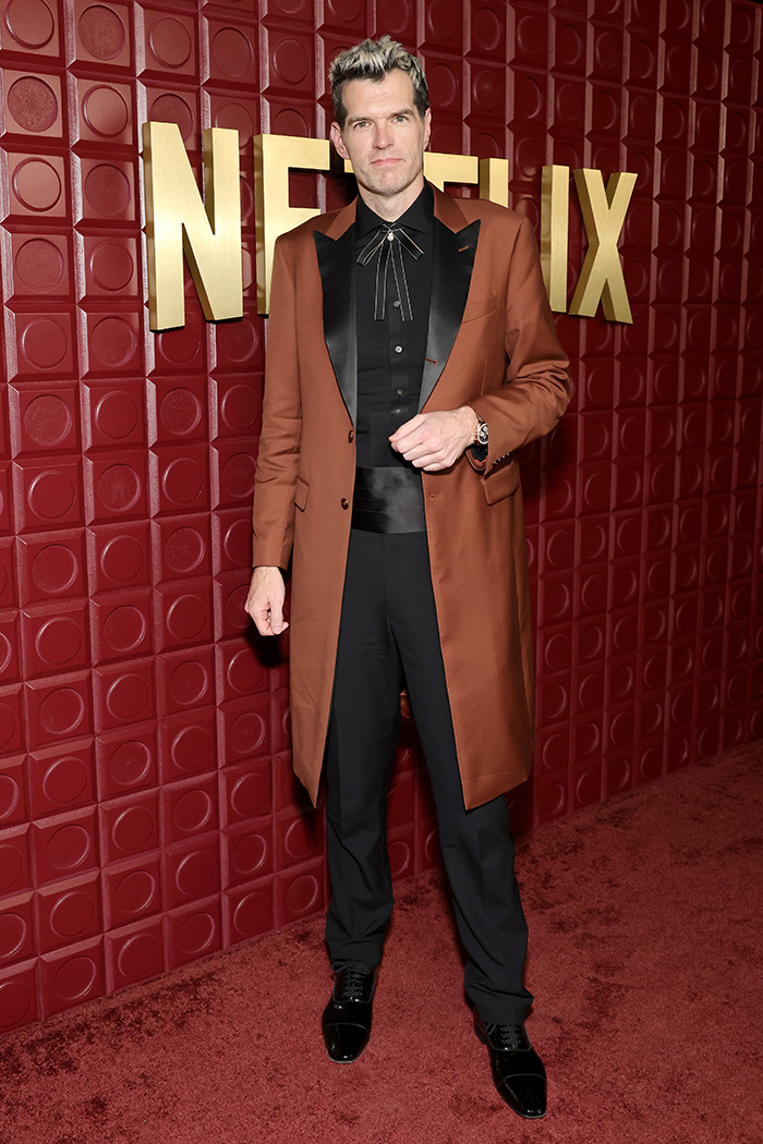 Male star in a brown coat and black outfit standing on a red carpet, representing worst dressed stars at Golden Globes after-parties.
