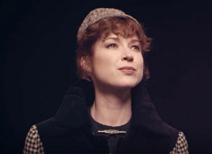 Woman with short curly hair wearing a vintage hat and coat, representing celebrity downfalls in a dramatic setting.