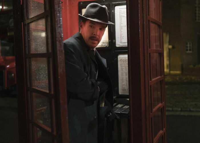 Actor in a vintage hat and coat inside a phone booth, depicting a scene about movie set workers and diva stars.