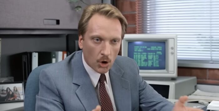 Man in a suit discussing pure evil villains, emphasizing Lord Voldemort, in an office with an old computer in the background.