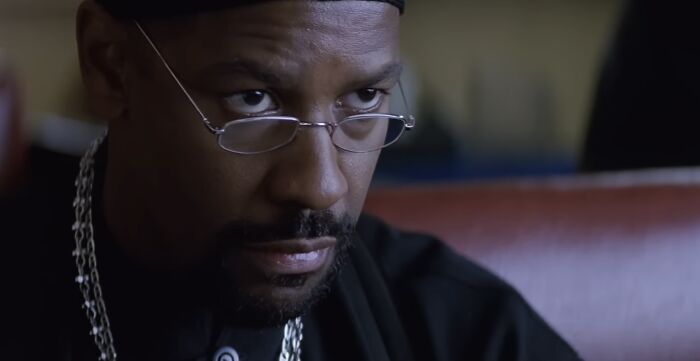 Close-up of a man with glasses and a chain necklace, portraying intensity in a dimly lit setting for villain analysis on Lord Voldemort.