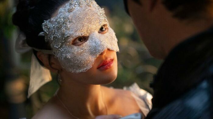 Actress behind Sophie Baek in Bridgerton wearing an ornate white lace mask during a close conversation scene. Actress behind Sophie Baek in Bridgerton wearing an ornate white lace mask during a close conversation scene.