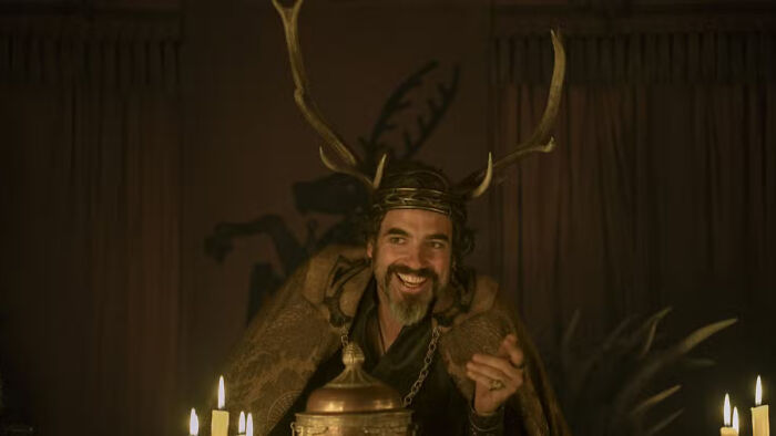 Man wearing antler crown and fur cloak in dimly lit room, representing a house sigil in Knight of the Seven Kingdoms.