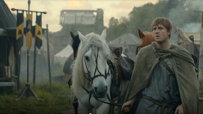 Medieval camp scene with a man leading a white horse, showcasing every house sigil in a Knight of the Seven Kingdoms setting.