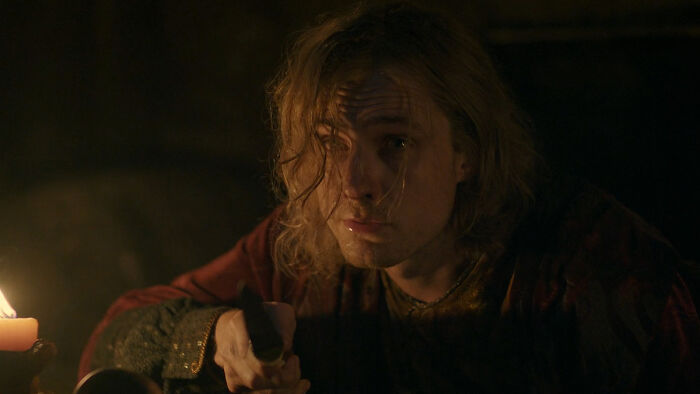 Targaryen actor from A Knight of the Seven Kingdoms holding a dagger with long hair in a dimly lit scene.