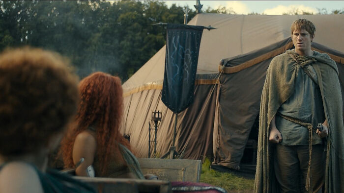 Medieval characters gathered near a large tent displaying house sigils from Knight of the Seven Kingdoms.