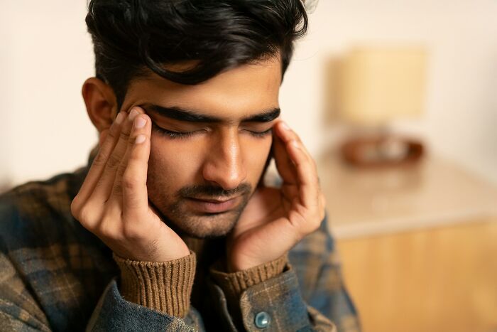 Young man with eyes closed and hands on temples, reflecting on adult goals and feeling overwhelmed indoors.