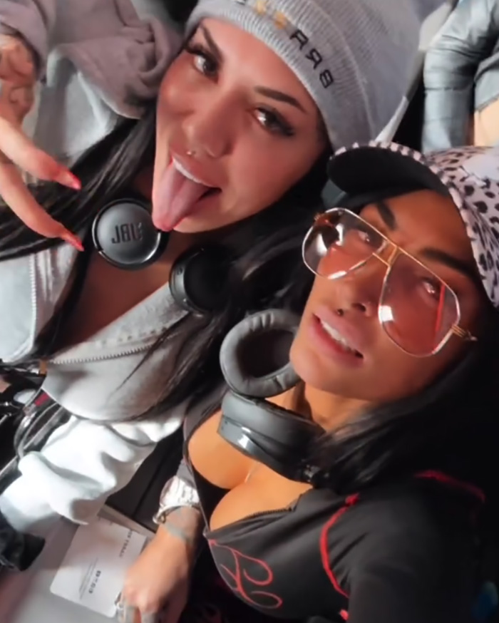 Two adult stars wearing headphones and casual outfits, posing for a selfie inside a plane first-class cabin.