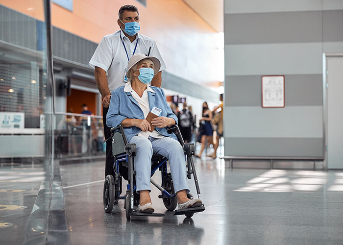 Elderly woman in wheelchair with a man pushing her, both wearing masks, at an airport during a heartbreaking goodbye.