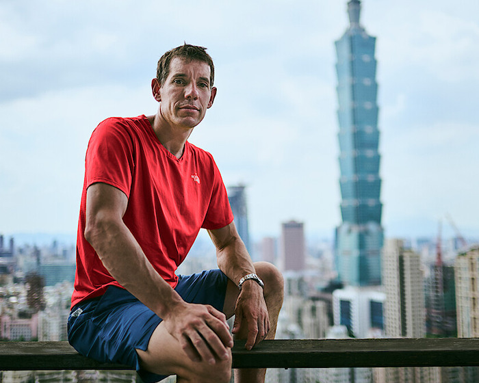 Famous climber in red shirt sitting with cityscape and skyscraper in the background, highlighting low pay controversy.