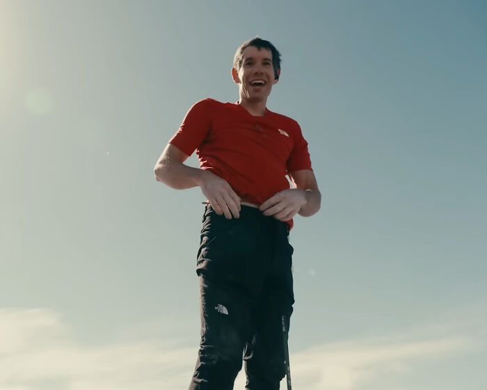 Famous climber smiling outdoors wearing a red shirt and black pants amid Netflix backlash over low pay for skyscraper climb.