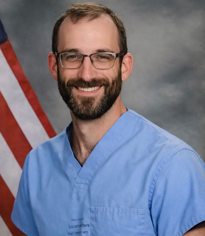 Man with glasses and beard wearing blue scrubs, smiling in front of an American flag, related to Alex Pretti&rsquo;s parents learning his fate.