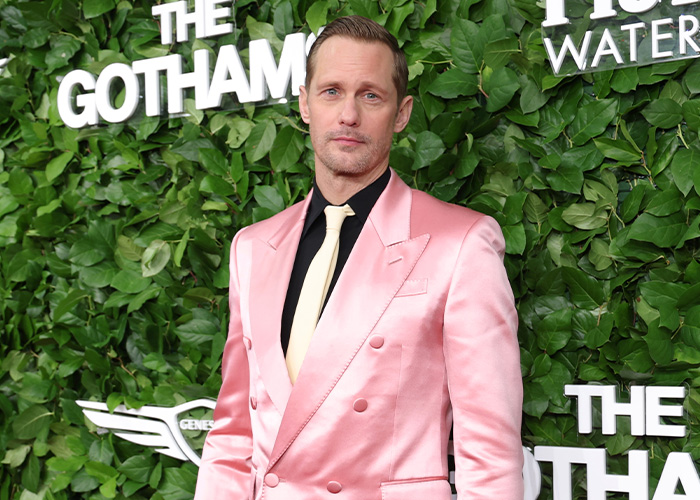 Alexander Skarsg&aring;rd posing at an event wearing a pink suit jacket with a black shirt and cream tie.
