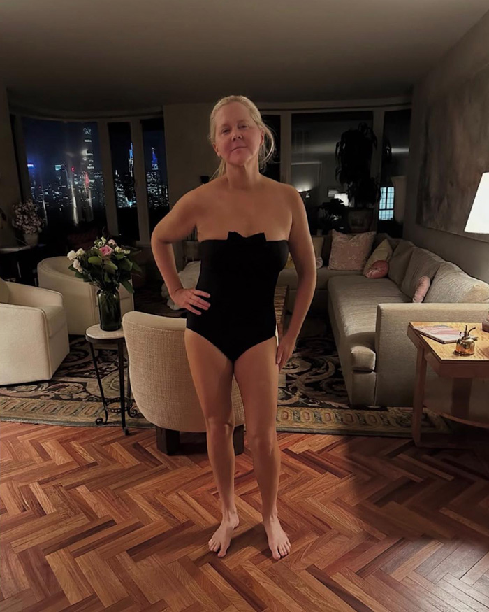 Amy Schumer in a black swimsuit standing in a living room with city lights visible through the windows at night.