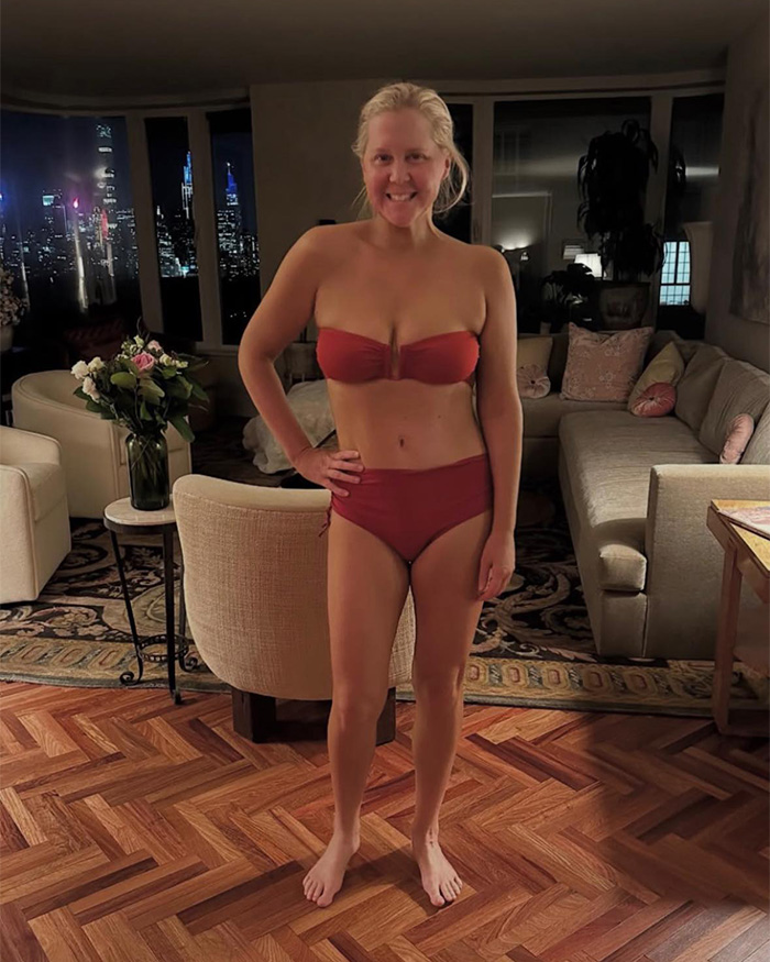 Amy Schumer smiling in a red bikini indoors with city lights visible through the window at night.