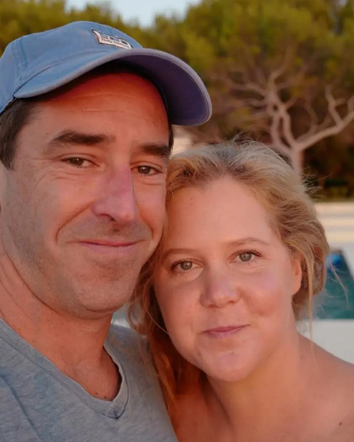 Amy Schumer and a man posing together outdoors, reflecting on her trolling herself after divorce filing.