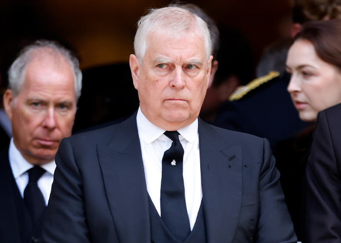 Prince Andrew in a black suit and tie, looking serious, with people in formal attire in the background.