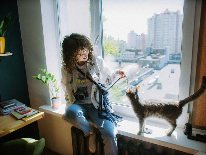 Woman sitting by a window playing with a cat, illustrating things women do to feel safer while living alone.