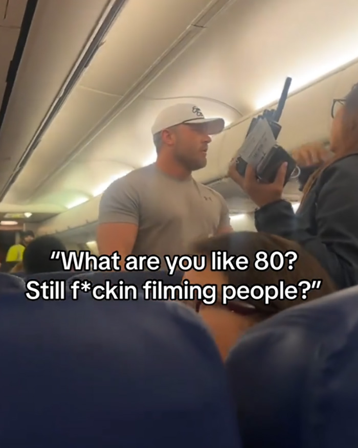 Bodybuilder in a gray shirt confronts elderly passenger on flight, showing anger before being removed by airline staff.