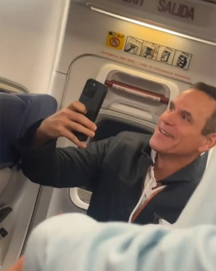 Man filming on phone inside airplane cabin during bodybuilder removal incident related to steroids controversy.