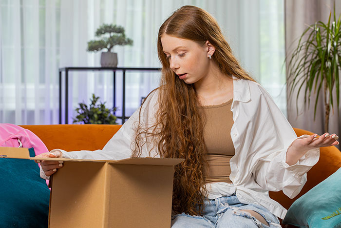Young woman with long hair sitting on a couch, looking confused while packing a cardboard box in a living room setting