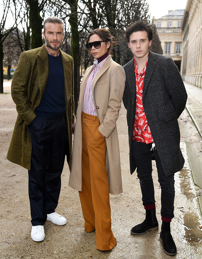 Victoria Beckham standing outdoors with two men, wearing sunglasses and a beige coat in a tense exchange amid feud.