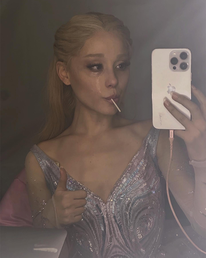 Actor in Wicked costume taking teary mirror selfie with lollipop and thumbs up in a dressing room
