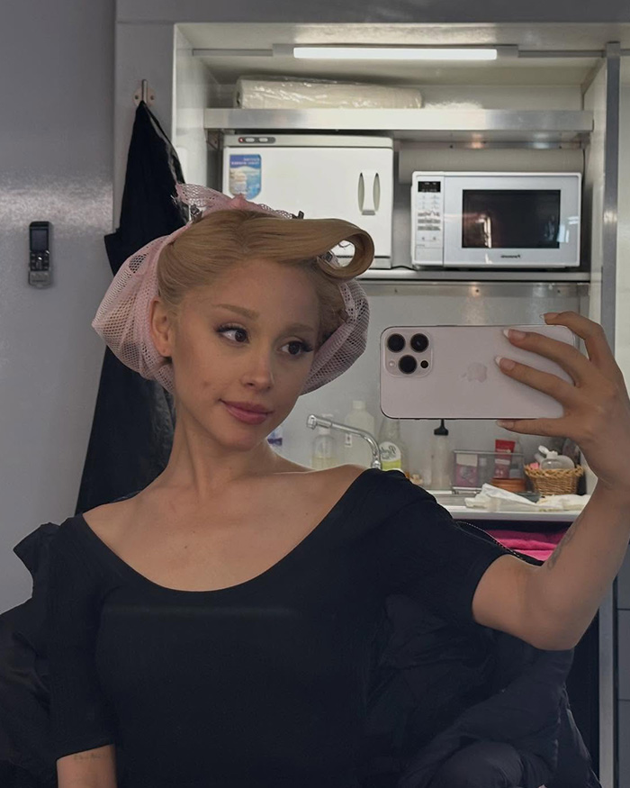 Performer mirror selfie in dressing room with curlers and hairnet, Wicked set photo showing makeup area and phone.