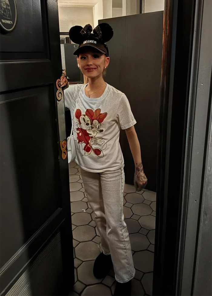 Young woman wearing a Mickey Mouse T-shirt and cap stands in a doorway, showcasing Ariana Grande's transformation in new pics.