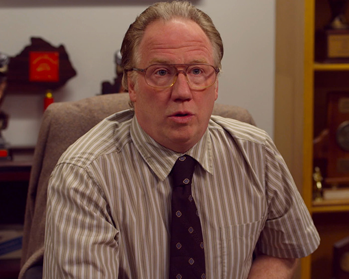 Middle-aged man in glasses and striped shirt sitting in an office, related to armed federal agents and Timothy Busfield.