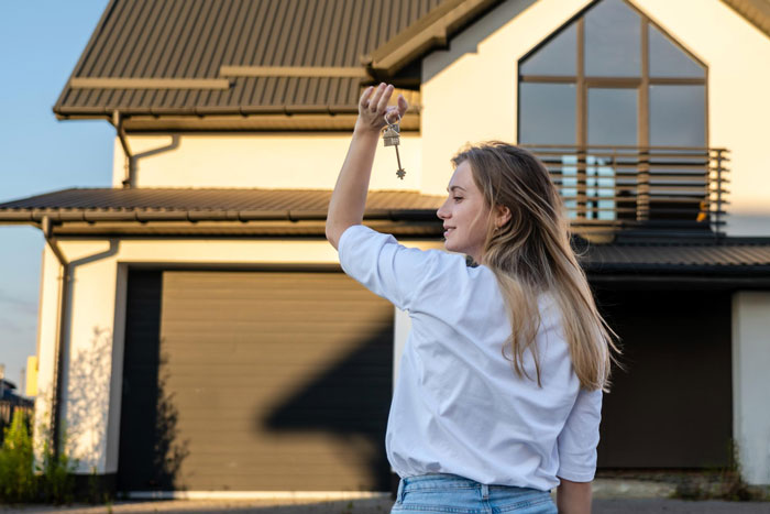 Young woman holding house keys in front of suburban home, parents house payment concept