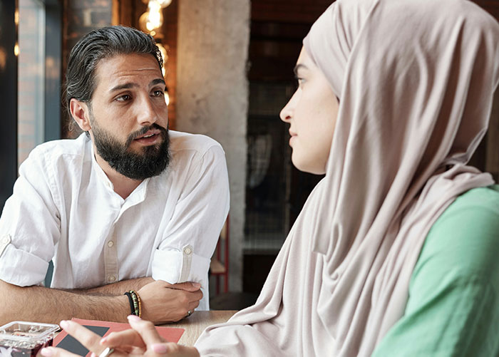 Man talking seriously to woman wearing hijab in a cafe, reflecting a guy refusing to pay for a hotel to help roommate.