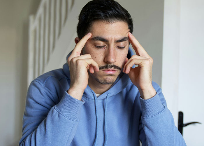 Young man in blue hoodie with eyes closed and fingers on temples appearing stressed about roommate and conservative family issues