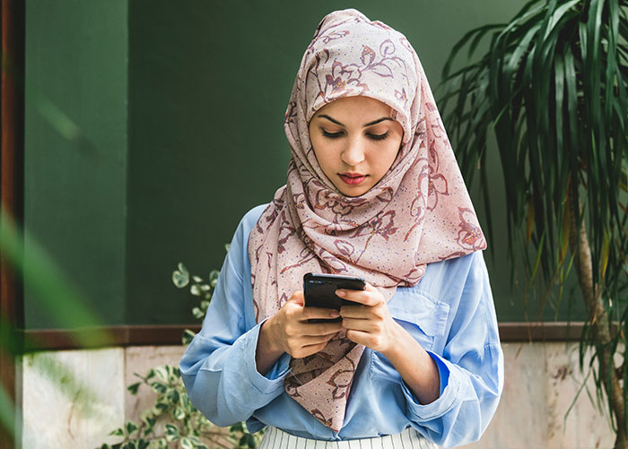 Young woman wearing a patterned hijab and blue shirt, focused on her smartphone, indoors with plants in the background
