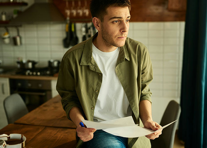 Young man in casual clothes holding papers, looking thoughtful while sitting in a modern kitchen setting.