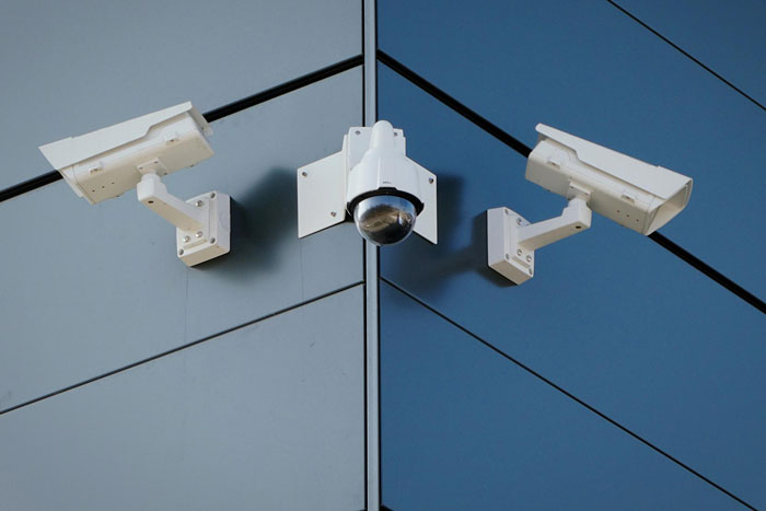 Three security cameras mounted on the corner of a modern glass building monitoring family safety and parole compliance. Three security cameras mounted on the corner of a modern glass building monitoring family safety and parole compliance.