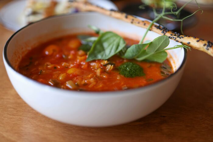 Bowl of tomato-based soup garnished with fresh herbs and served with a sesame breadstick on wooden table.