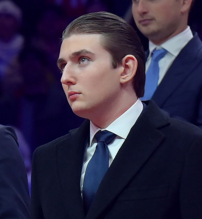 Barron Trump wearing a black coat and blue tie, attending an event with a focused expression in dim lighting.
