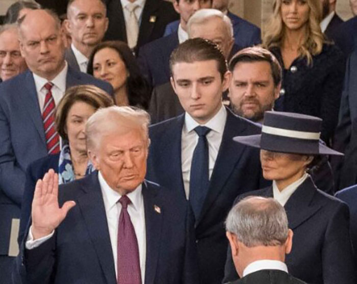 Barron Trump standing behind Donald Trump during a formal event, appearing serious and attentive among a crowd.