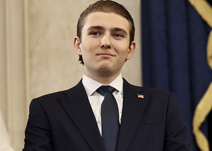 Barron Trump in a dark suit and tie, posing formally indoors, linked to paternity conspiracy theories after NYE event. Barron Trump in a dark suit and tie, posing formally indoors, linked to paternity conspiracy theories after NYE event.
