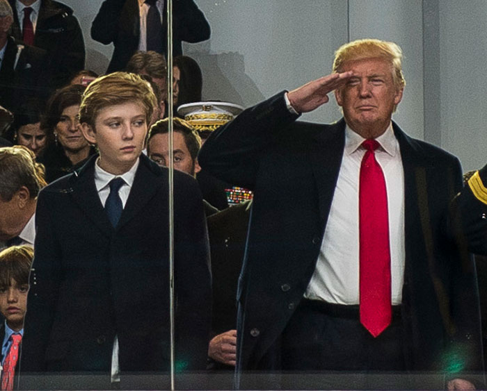 Barron Trump standing beside a man saluting during a formal event with people seated in the background.