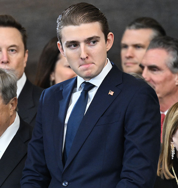 Barron Trump in a navy suit looking concerned at a formal event with blurred people in the background.